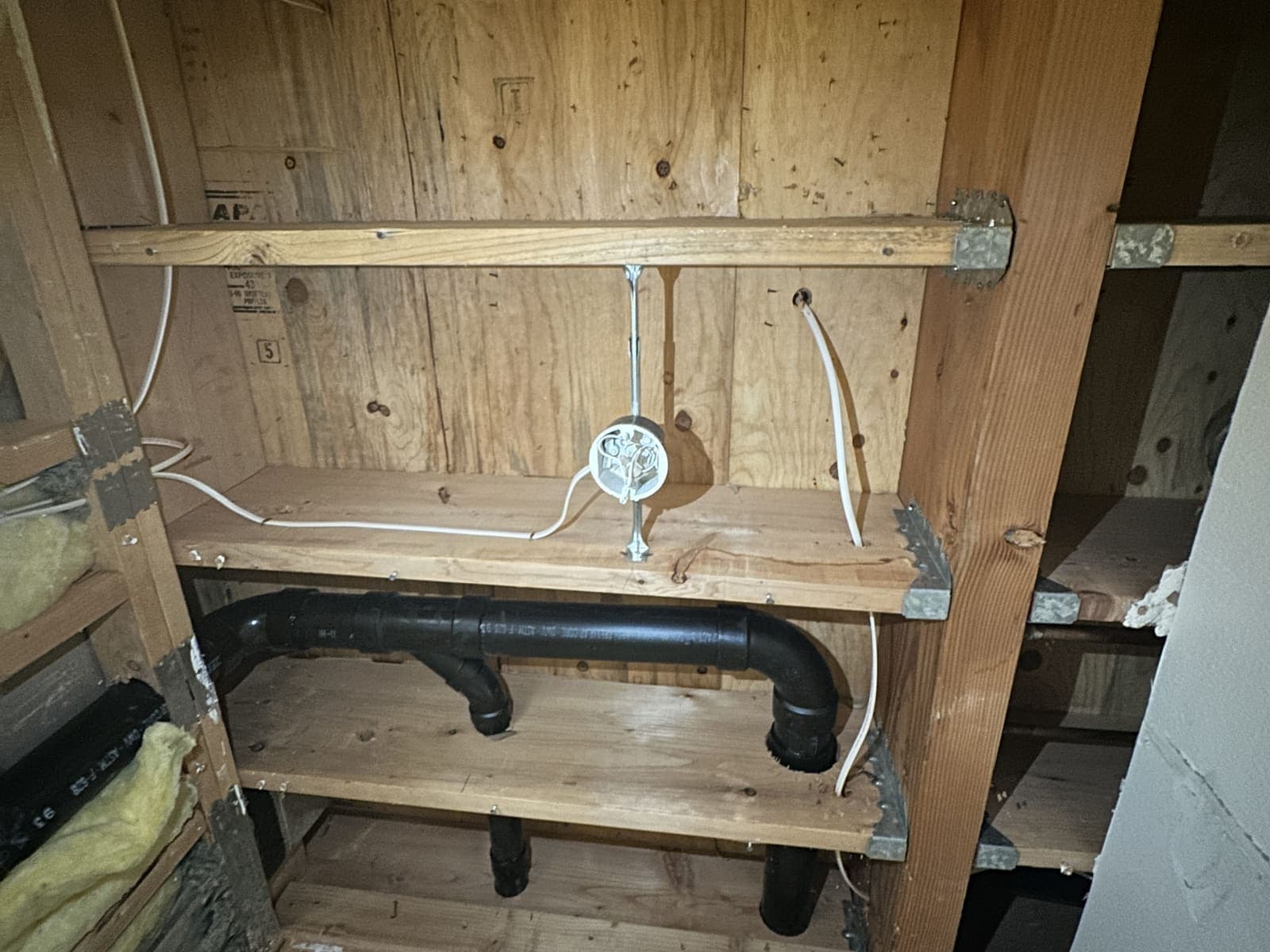 An opened wall and ceiling bay showing a black drain line, electrical wiring and a junction box running between the framing