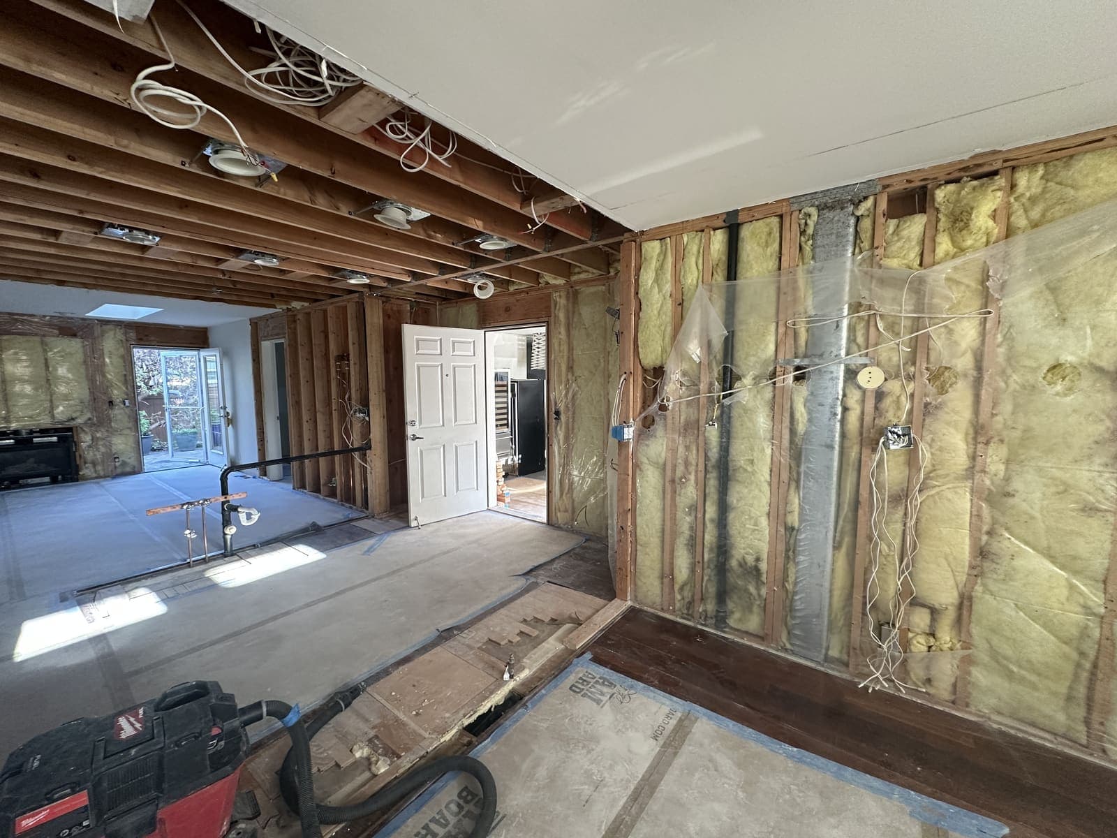 A kitchen mid-demolition: open ceiling joists, loose wiring, insulated stud walls and plumbing stubbed up through the subfloor