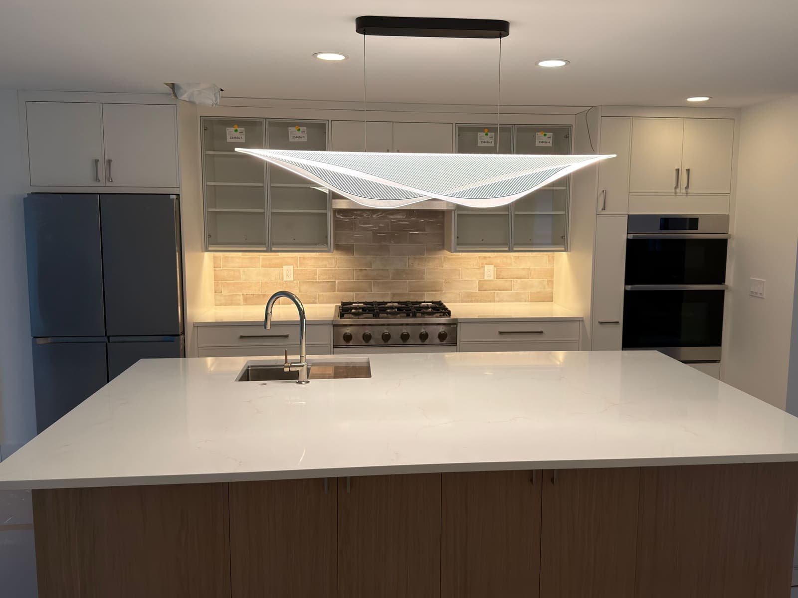The same kitchen after the remodel: white cabinets with glass-front uppers, a tile backsplash, a gas range and a large island under a pendant light