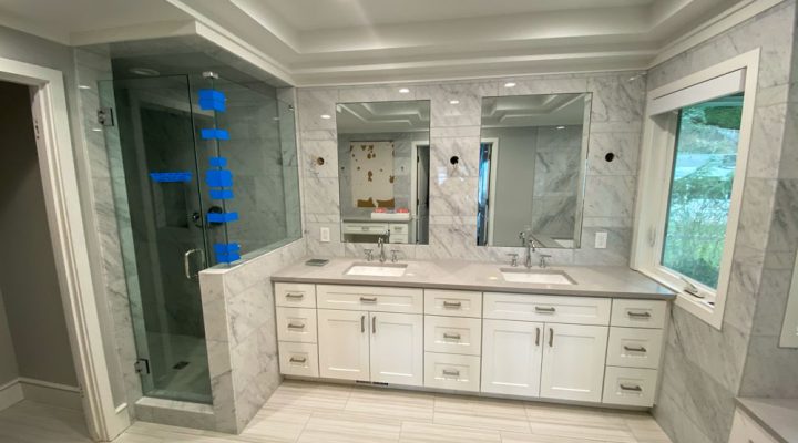 A remodeled modern bathroom featuring a double vanity with two faucets and a spacious walk-in shower with a glass enclosure. Light enters the bathroom through a window near the shower.