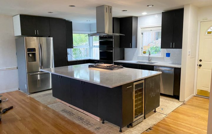 A large, open-plan kitchen with a central island that doubles as a breakfast bar. The island has a porcelain countertop and stainless steel sink, and is lit by recessed lights. Stainless steel appliances line the back wall, and dark wood cabinets with metal handles run along both sides of the kitchen.
