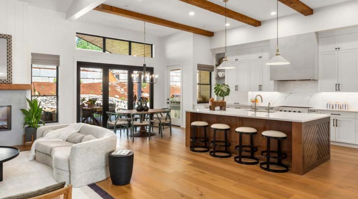 A large, open kitchen with a vaulted ceiling. Exposed wooden beams run across the ceiling. Light colored wooden cabinets line the back wall, with a stainless steel sink and faucet in the center. The dining area is on the left with a view of the outdoor through the glass doors.